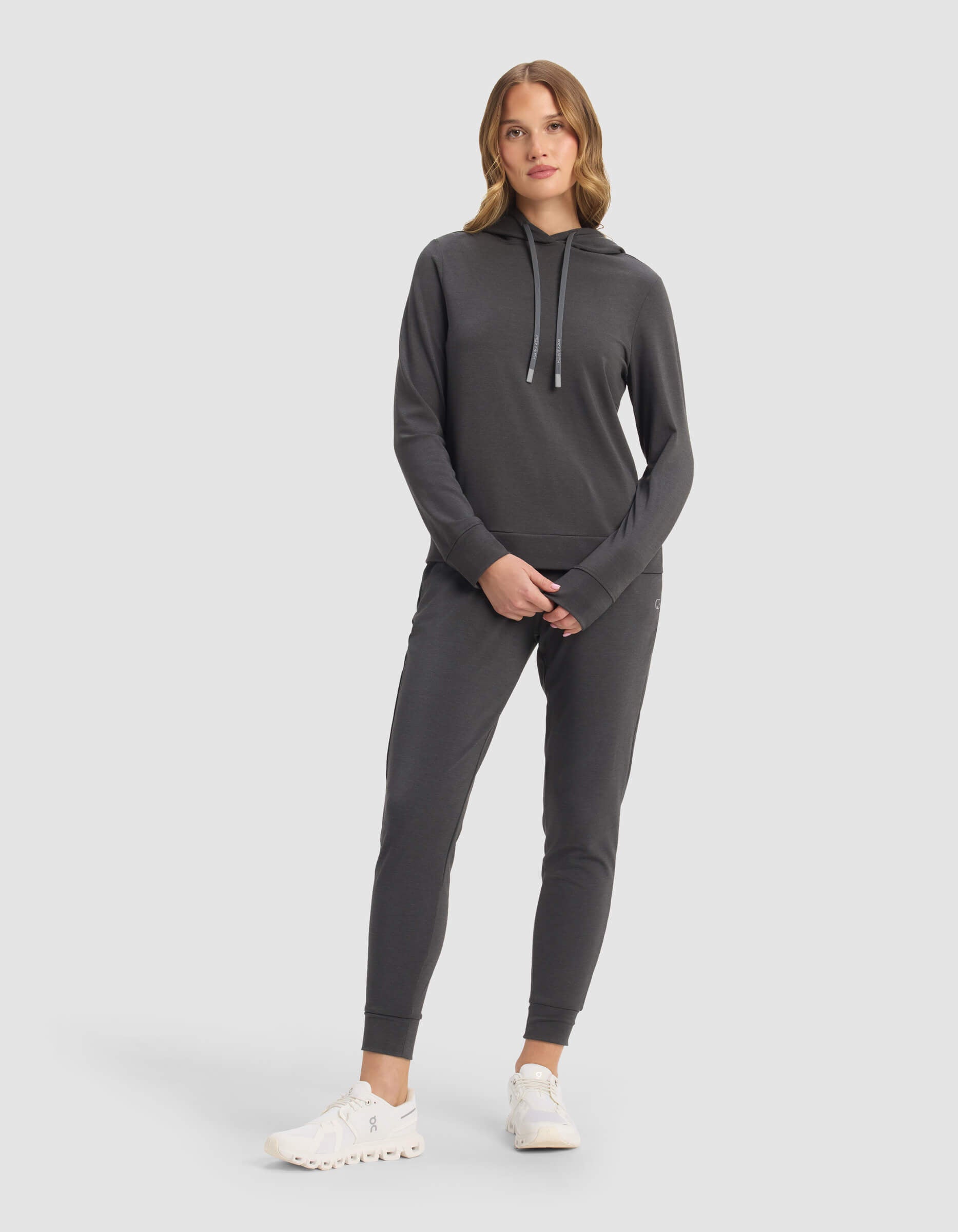 A woman with light brown hair stands against a plain background, hands in pockets, wearing the Cozy Earth Women's Brushed Bamboo Hoodie & Jogger Set in dark grey, paired with white athletic shoes.