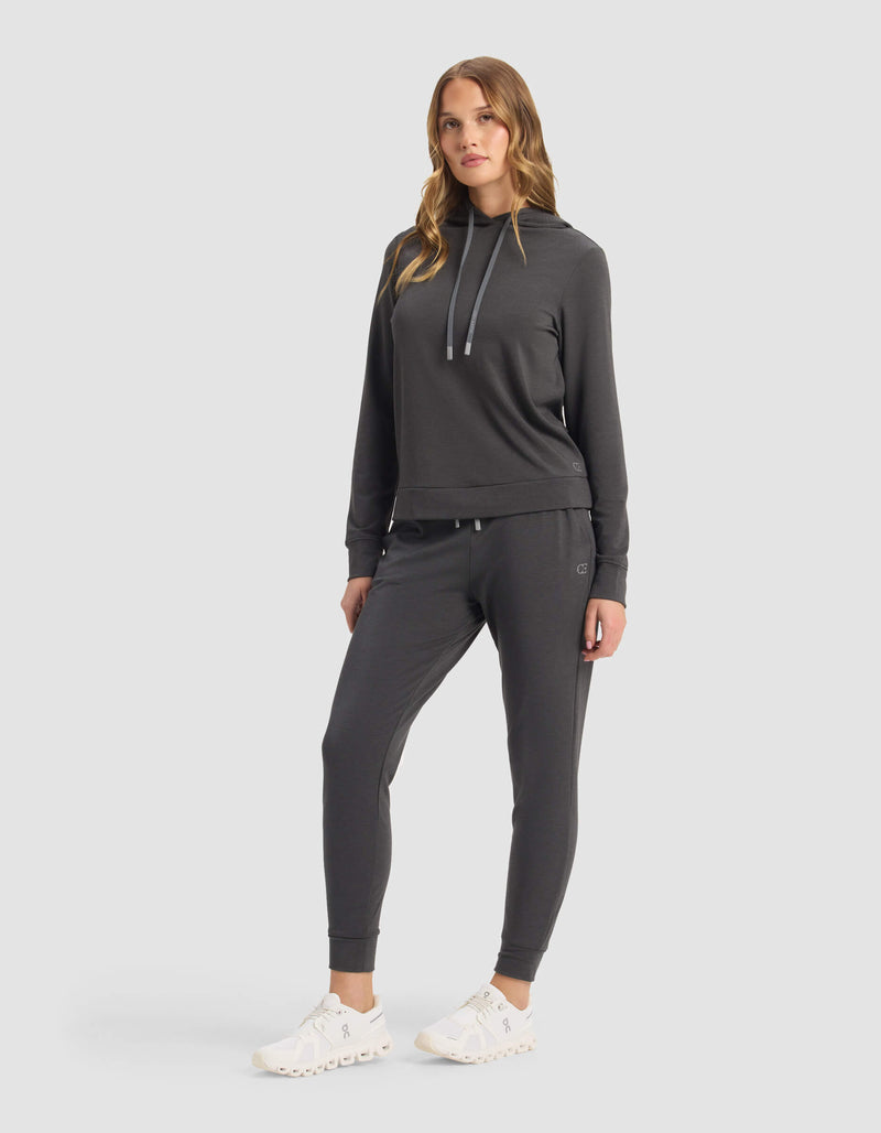 A woman in a relaxed pose stands against a light gray background, wearing the Cozy Earth Women's Brushed Bamboo Hoodie & Jogger Set in dark gray, paired with white sneakers. She has long, wavy hair with one leg slightly forward.