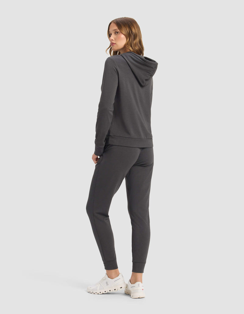 A person faces away, glancing over their shoulder in a Cozy Earth Women's Brushed Bamboo Hoodie & Jogger Set, dark gray. White slip-on shoes and a plain light gray background complete the look.