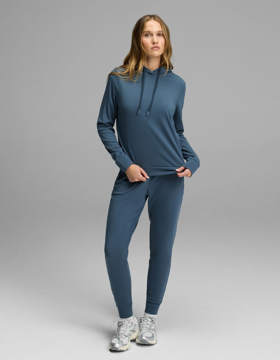 A woman with long blonde hair stands on a gray backdrop wearing the Cozy Earth Women’s Brushed Bamboo Hoodie & Jogger Set in blue, paired with white running shoes. She is holding part of her sweatshirt.|Color:Dawn