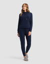 A woman with long, wavy hair stands against a plain background wearing the Cozy Earth Women's Brushed Bamboo Hoodie & Jogger Set in navy blue, paired with gray slippers, looking slightly to the side.|Color:Navy