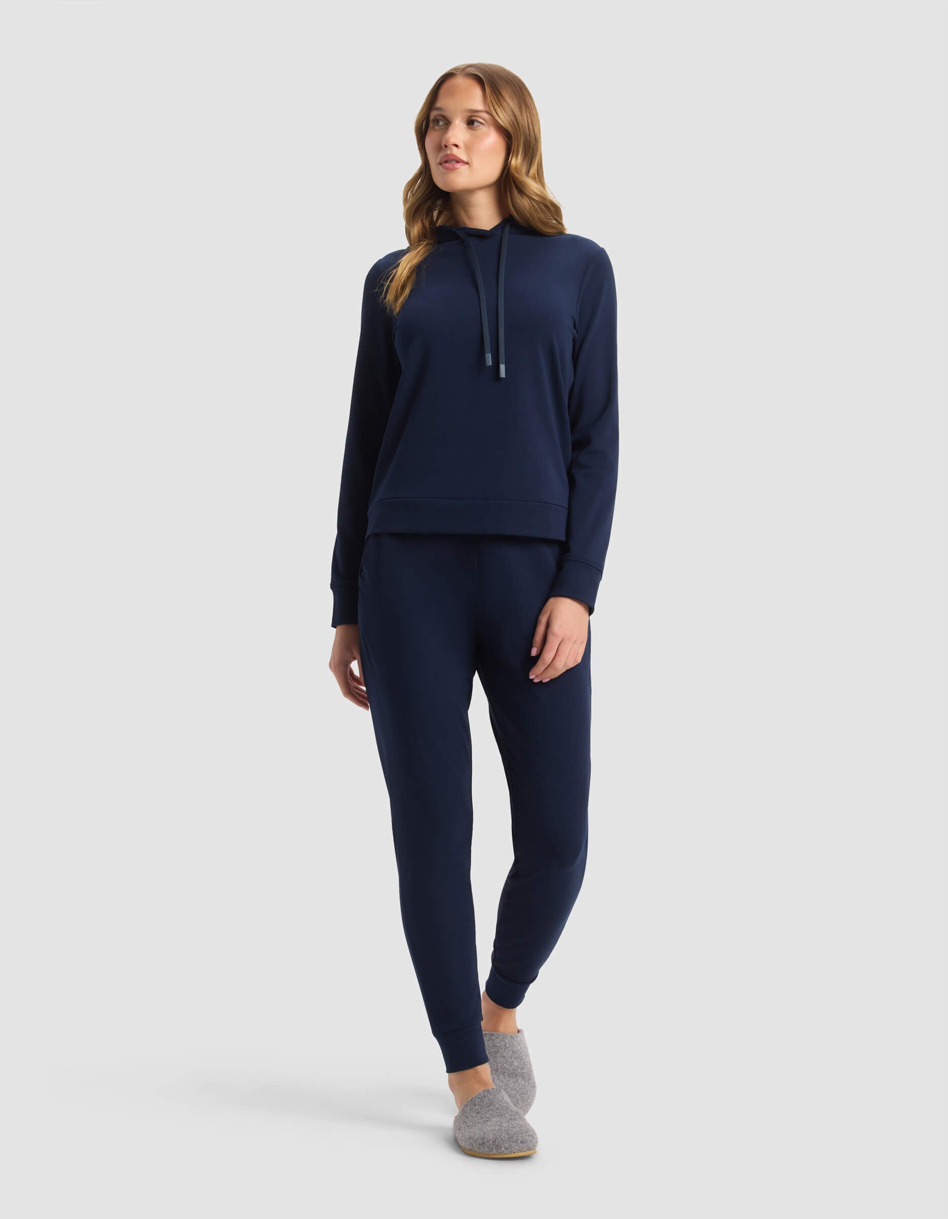 A woman with long, wavy hair stands against a plain background wearing the Cozy Earth Women's Brushed Bamboo Hoodie & Jogger Set in navy blue, paired with gray slippers, looking slightly to the side.