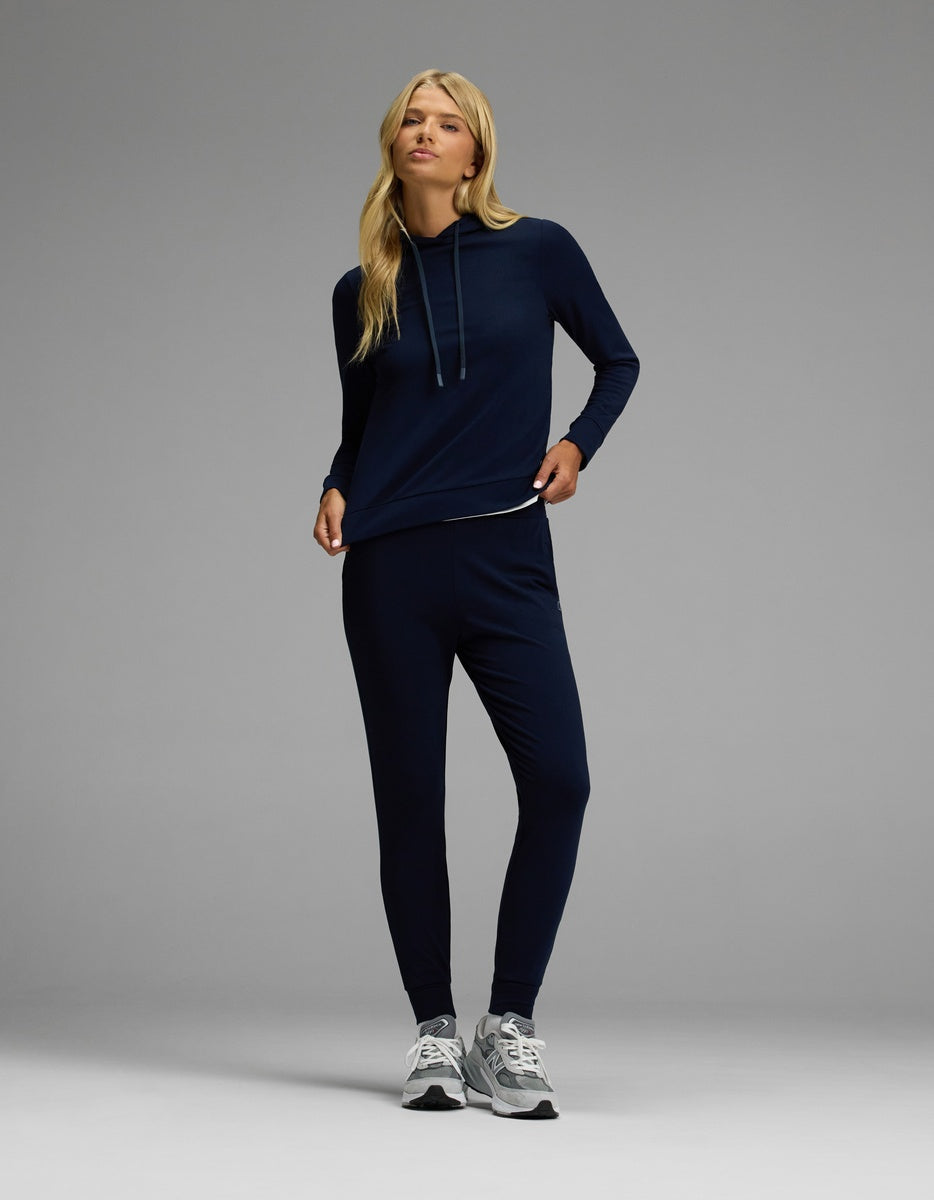 A model wears the Cozy Earth Women’s Brushed Bamboo Hoodie & Jogger Set in navy blue with gray sneakers, standing against a plain gray background and glancing to the side while holding the hem of the hoodie.|Color:Navy