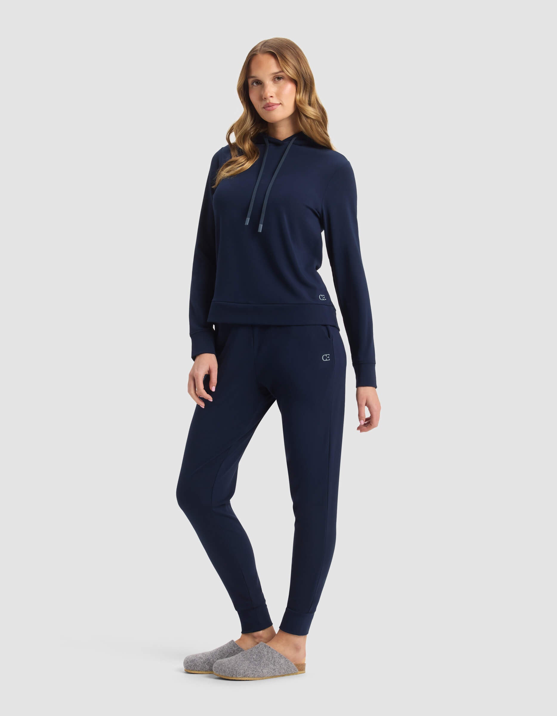 A woman poses in a relaxed stance wearing the Cozy Earth Women's Brushed Bamboo Hoodie & Jogger Set in navy, paired with light gray slippers, set against a plain light gray background.