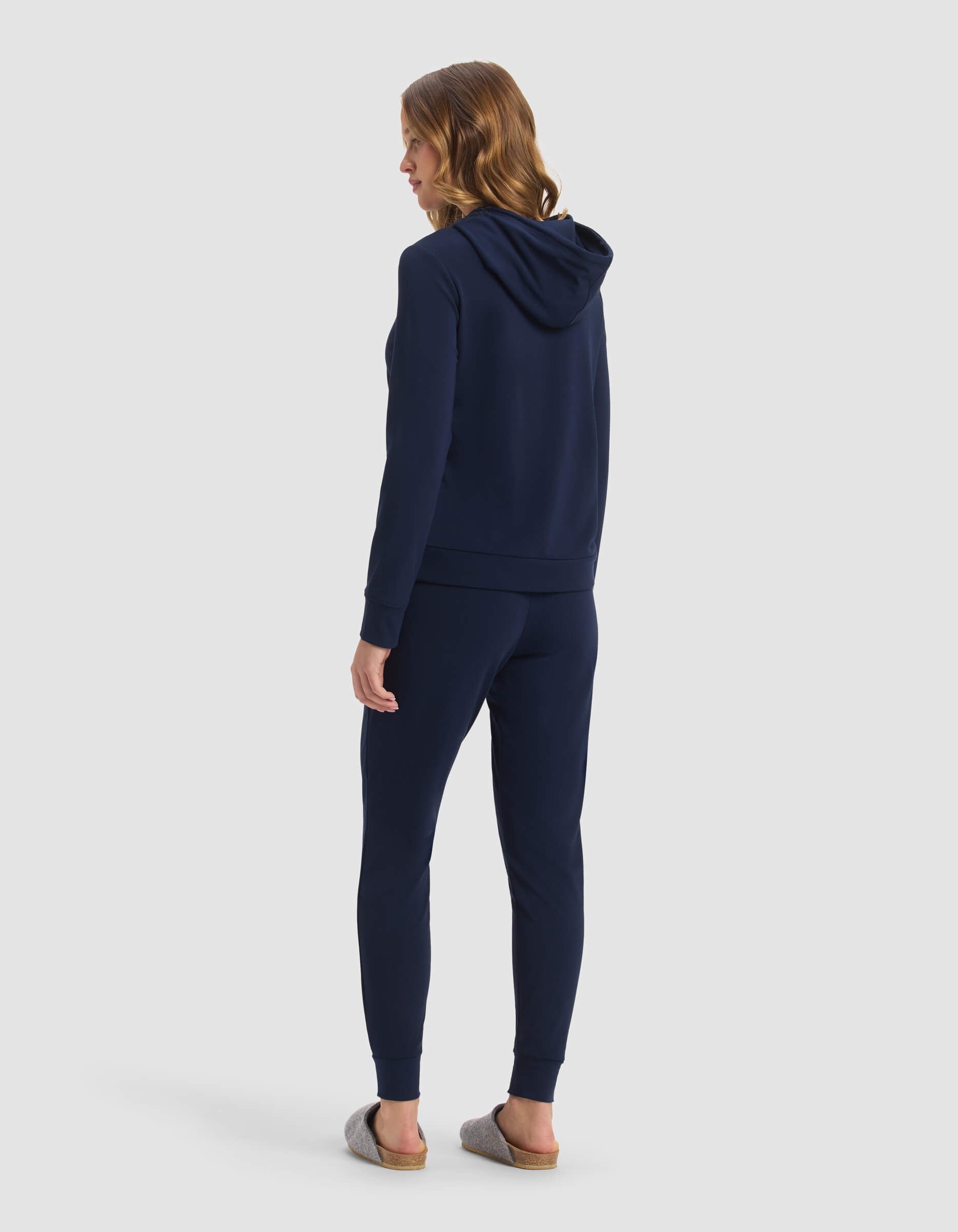 A woman faces away wearing the Cozy Earth Women's Brushed Bamboo Hoodie & Jogger Set in navy, paired with gray slip-on shoes, set against a plain light background.|Color:Navy