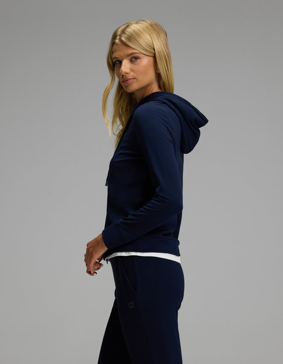 A woman with long blonde hair is wearing the Cozy Earth Women’s Brushed Bamboo Hoodie & Jogger Set in navy blue, standing sideways and looking at the camera against a neutral grey background.
