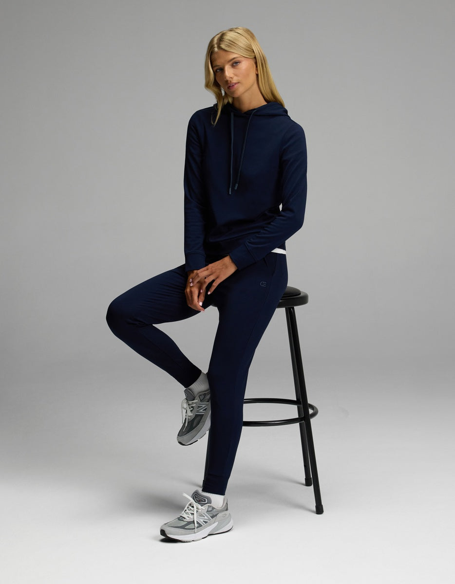 A woman wears the Cozy Earth Women’s Brushed Bamboo Hoodie & Jogger Set in navy as she sits on a black stool with one leg up, pairing the outfit with gray sneakers against a plain gray background.
