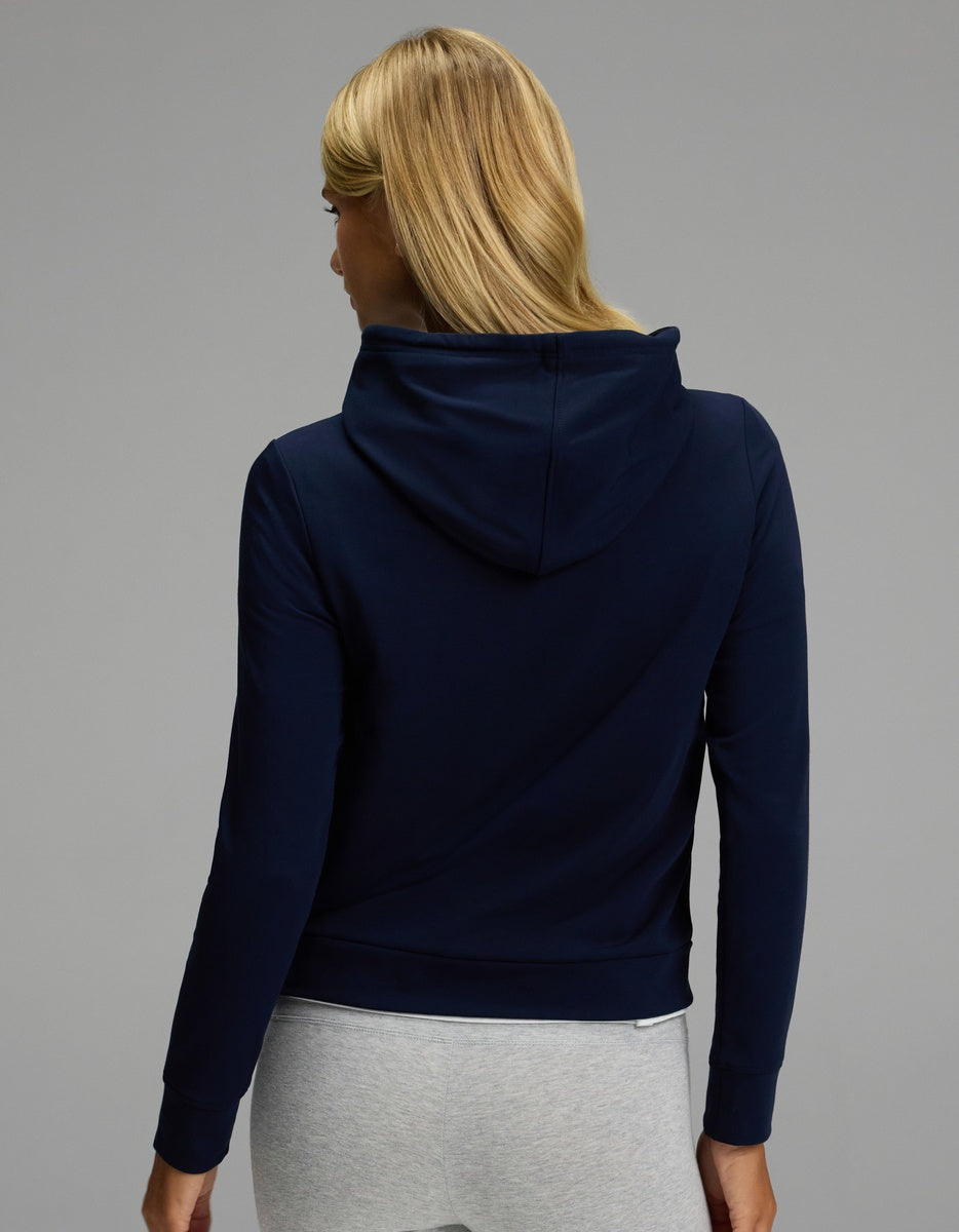 A person with long blonde hair models the Cozy Earth Women’s Brushed Bamboo Hoodie in navy, paired with light gray pants, facing away from the camera against a plain gray background.