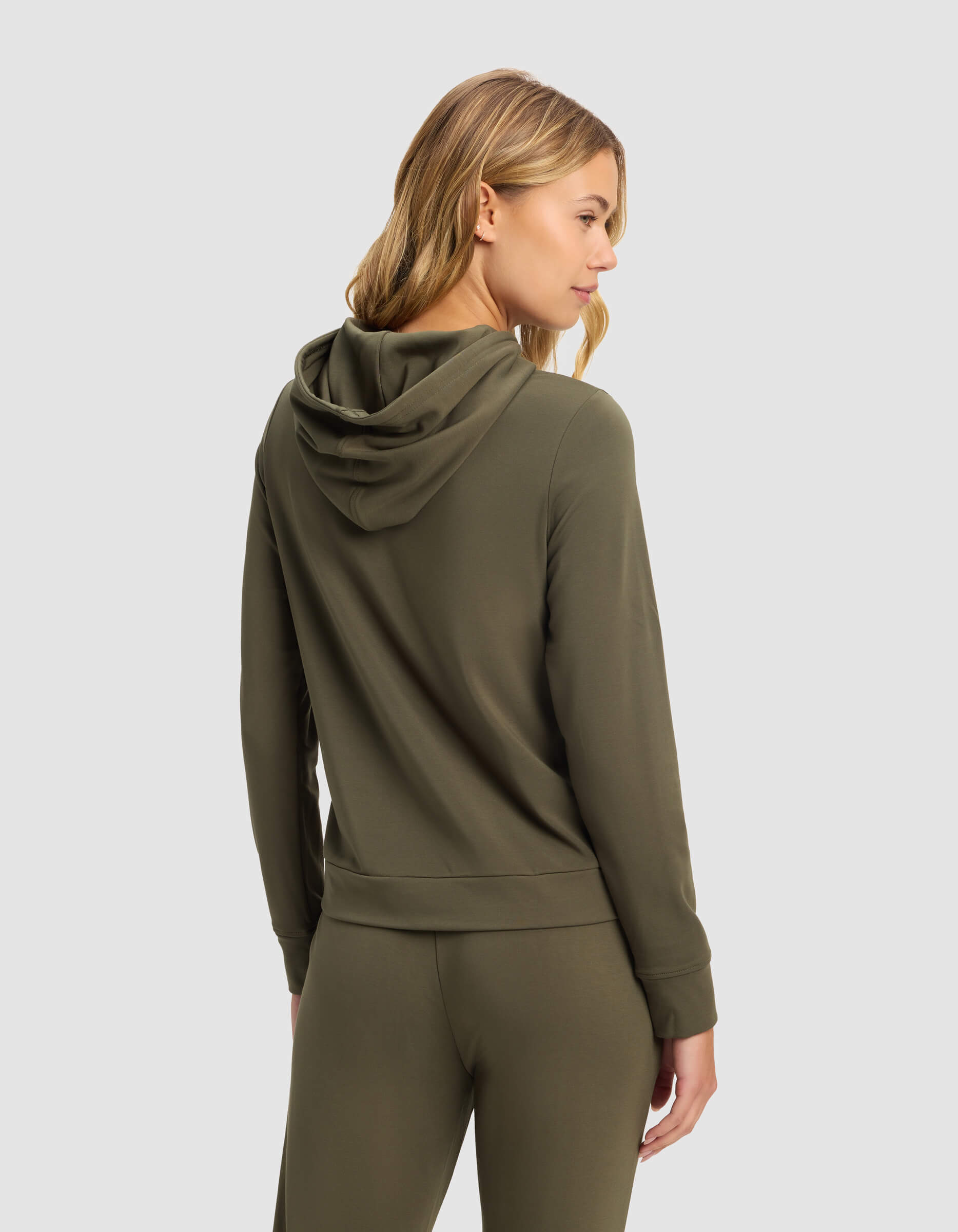 A woman with blonde hair models the Cozy Earth Women's Brushed Bamboo Hoodie in olive green, paired with matching pants, standing with her back and head slightly turned against a plain light gray background.