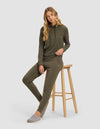 A woman with long blonde hair models the Cozy Earth Women's Brushed Bamboo Hoodie in olive green, paired with gray slippers, while sitting on a wooden stool against a plain white background.|Color:Olive