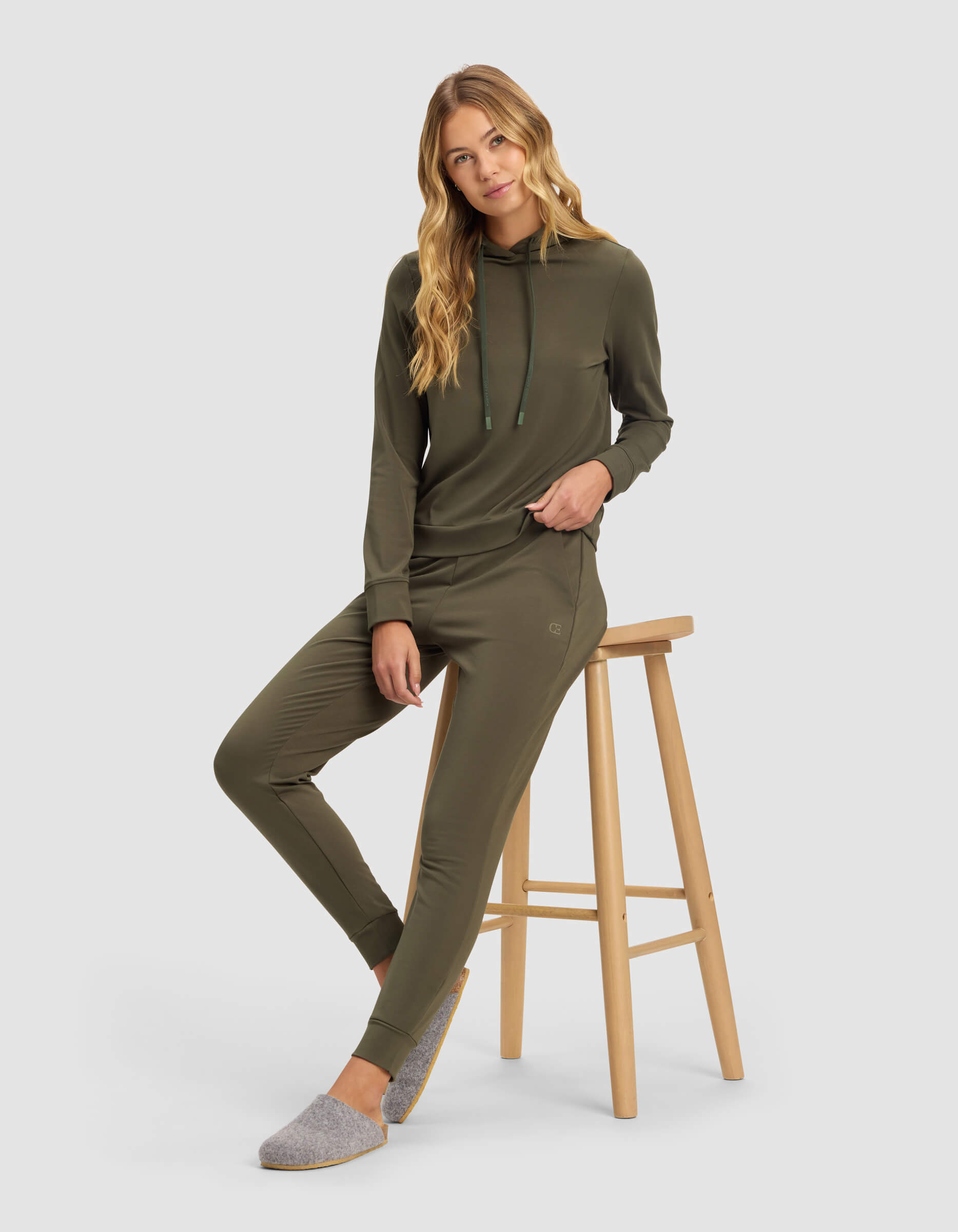 A woman with long blonde hair models the Cozy Earth Women's Brushed Bamboo Hoodie in olive green, paired with gray slippers, while sitting on a wooden stool against a plain white background.