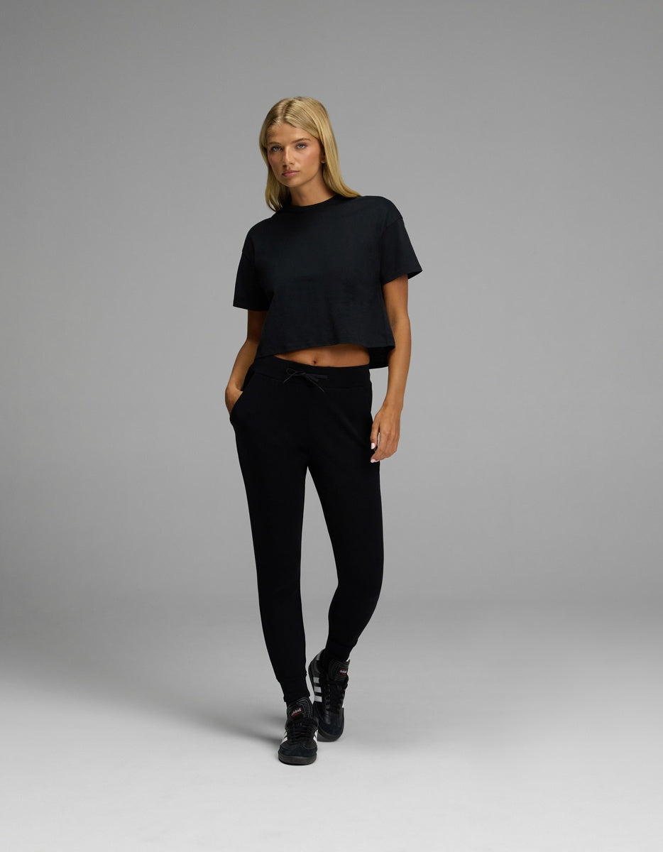 A person with long blonde hair stands with one foot forward and hands in pockets against a plain gray background, wearing black sneakers, a black cropped t-shirt, and Cozy Earth’s Women’s Brushed Bamboo Jogger.