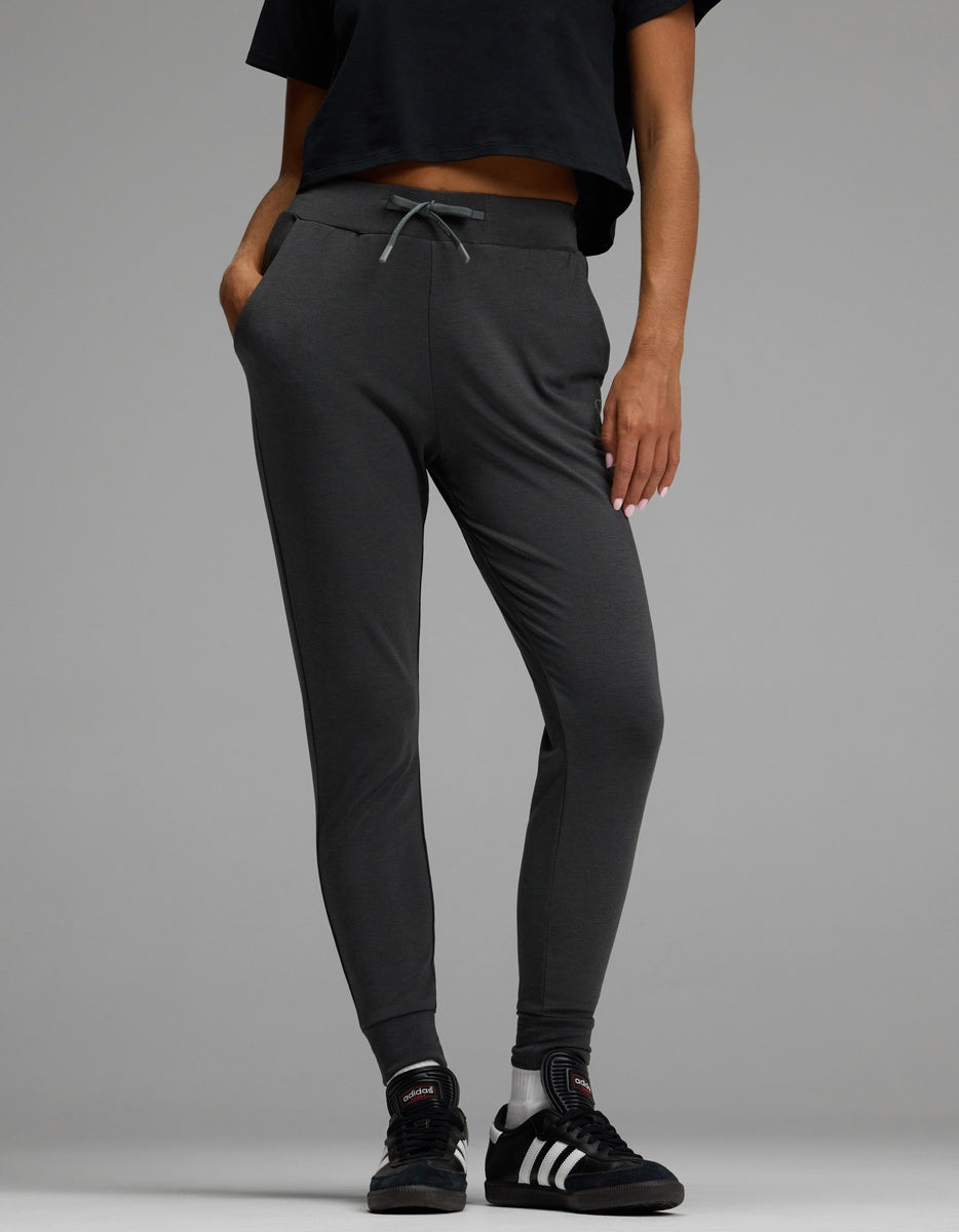 A person wearing Cozy Earth’s Women’s Brushed Bamboo Jogger in dark gray, paired with a black cropped shirt and black Adidas sneakers with white stripes, stands with hands in their pockets against a plain gray background.|Color:Charcoal
