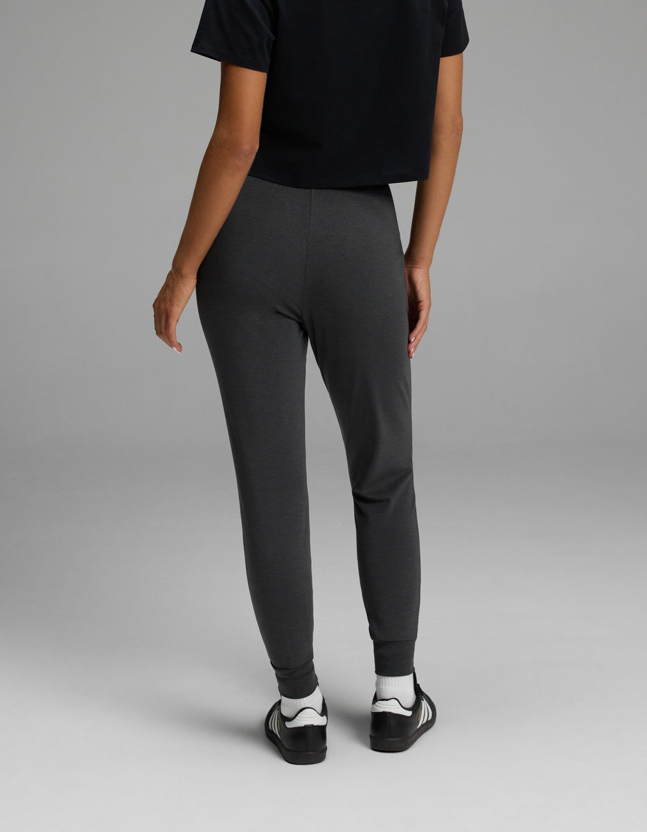 A person is standing on a gray floor, facing away, wearing a black T-shirt, black-and-white sneakers, and Cozy Earth Women’s Brushed Bamboo Jogger pants in dark gray. Only the lower back, legs, and part of the arm are visible.