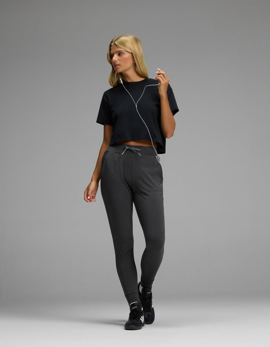 A woman with blonde hair is standing against a plain gray background, looking to the side while listening to earphones. She is wearing a black cropped t-shirt, black sneakers, and the Cozy Earth Women’s Brushed Bamboo Jogger in dark gray.