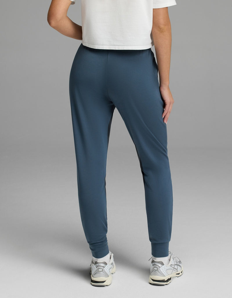 A person is seen from the back, wearing a white cropped T-shirt, white sneakers, and Cozy Earth’s Women’s Brushed Bamboo Jogger in blue, standing against a plain gray background.