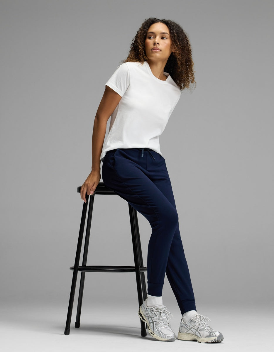 A woman wearing Cozy Earth’s Women’s Brushed Bamboo Jogger in navy, paired with a white t-shirt and gray sneakers, sits sideways on a tall black stool against a plain gray background, looking to her left.