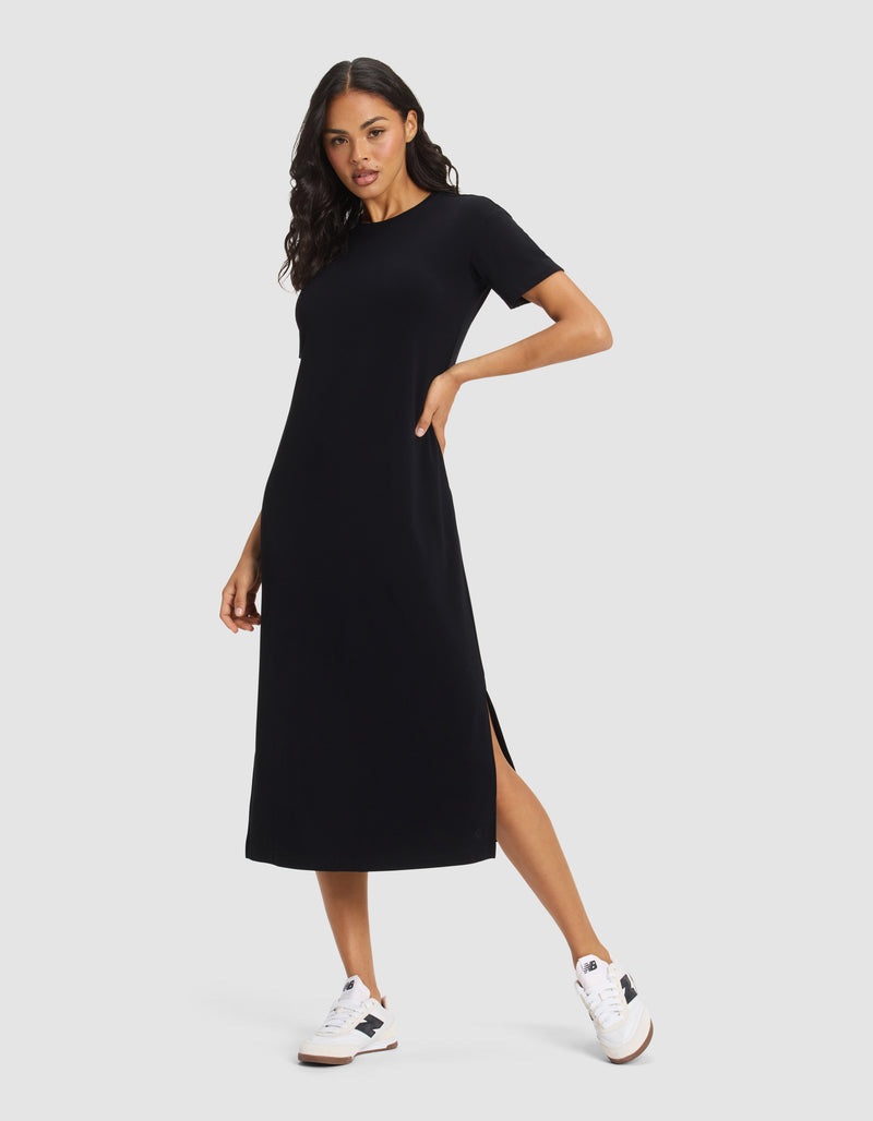 A woman models the Cozy Earth Women's Brushed Bamboo Midi Dress—short-sleeve, black, ankle-length with side slit—paired with white sneakers against a plain light grey background.