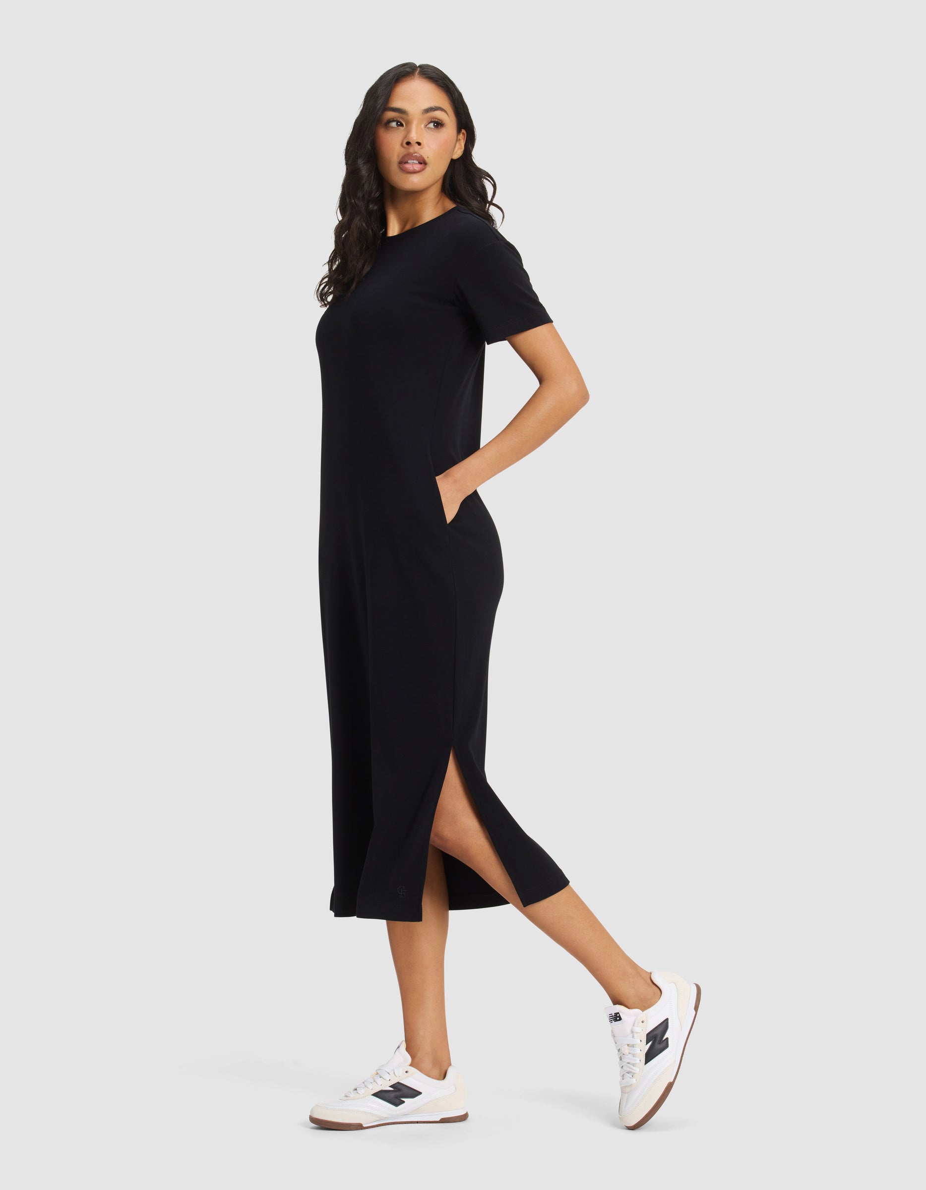 A woman stands sideways wearing the Cozy Earth Women's Brushed Bamboo Midi Dress with short sleeves, side slits, and pockets, paired with white sneakers. She looks ahead neutrally against a plain background.|Color:Black