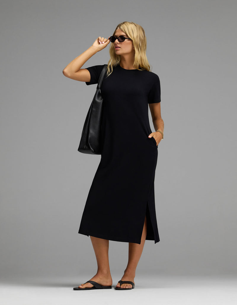 A woman stands confidently against a plain gray background, wearing the Cozy Earth Women’s Brushed Bamboo Midi Dress in black with short sleeves and side slits. She completes her look with black sandals, sunglasses, and a large black tote bag.