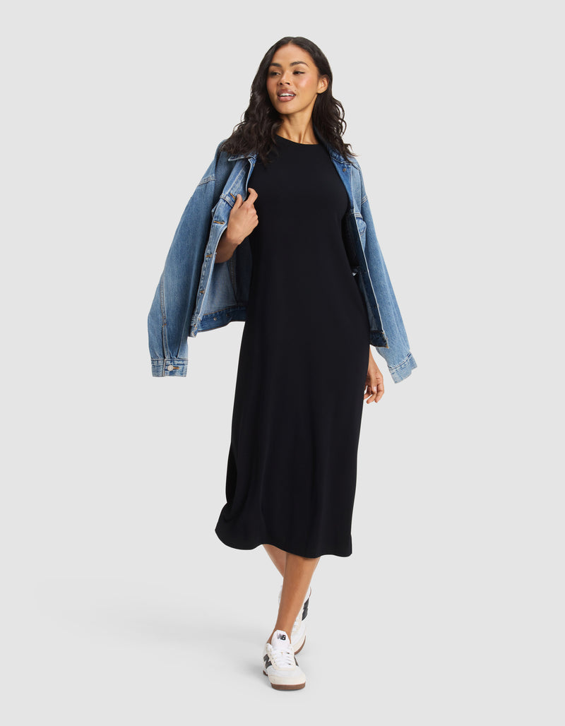 A woman poses against a plain light background wearing Cozy Earth's Women's Brushed Bamboo Midi Dress in black, paired with white sneakers and a blue denim jacket draped over her shoulders.