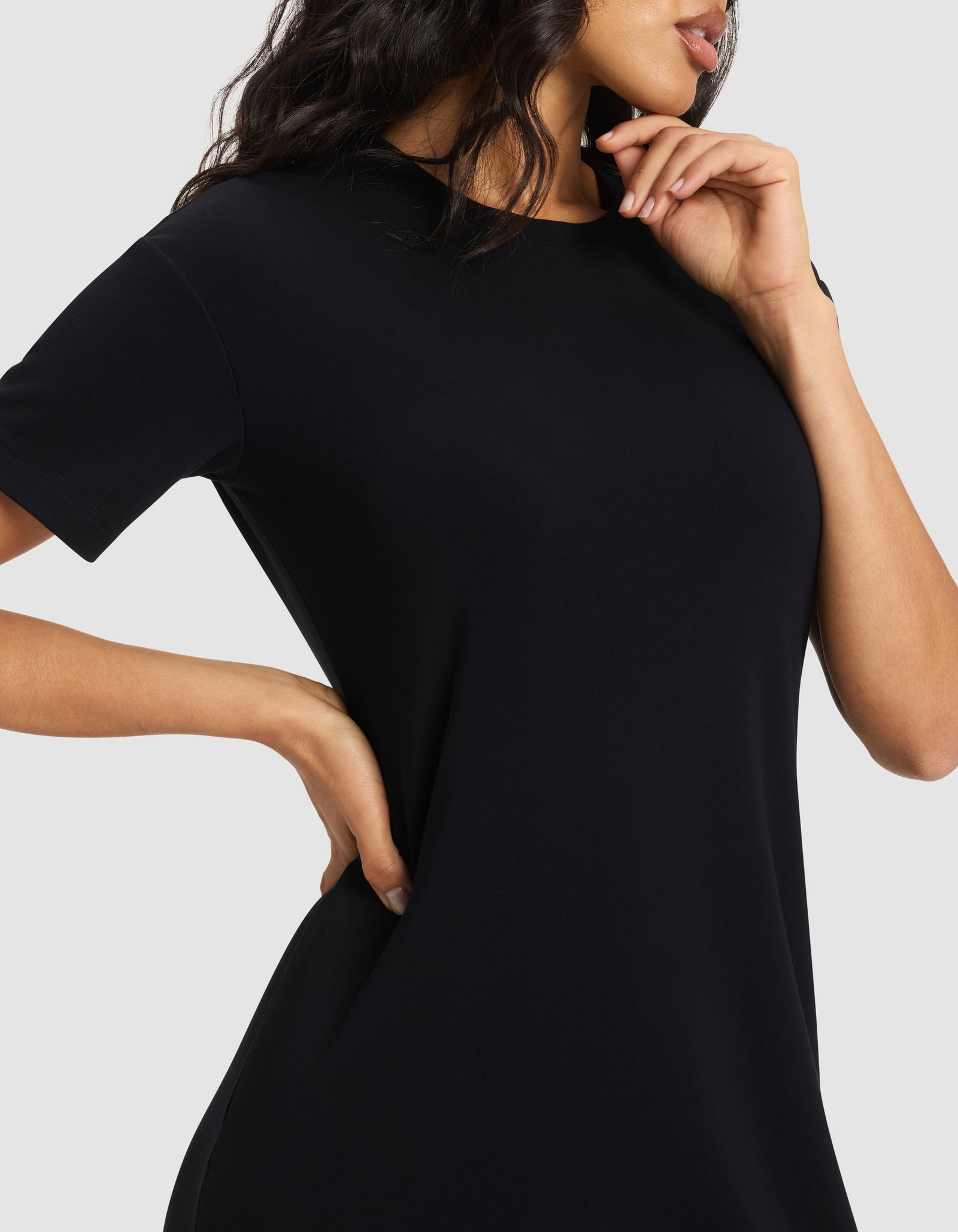 A woman models the Cozy Earth Women's Brushed Bamboo Midi Dress, a short-sleeved black style, posing with one hand on her hip and the other near her face against a plain light gray background. Her face is partially visible.