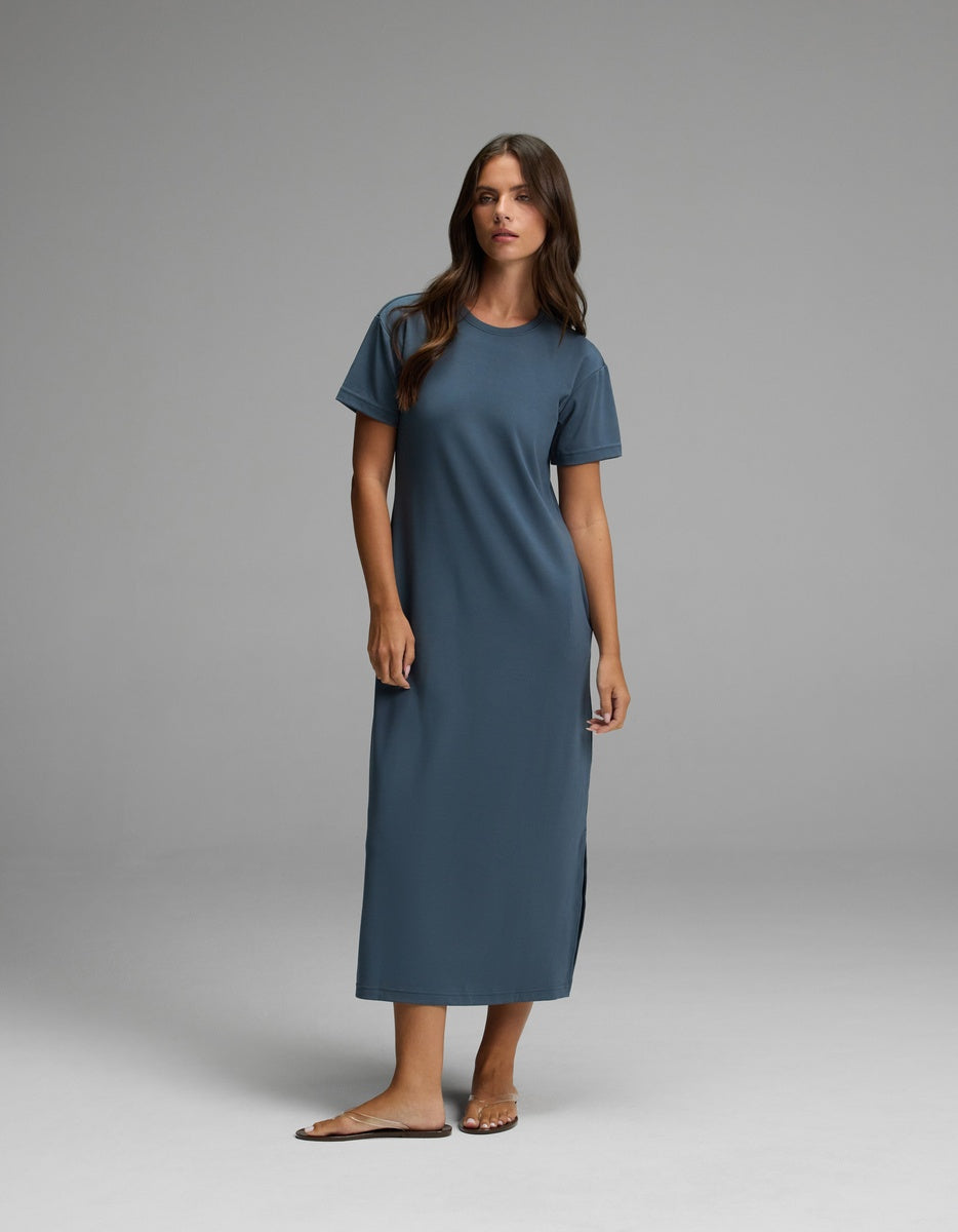 A woman stands in a studio against a plain gray background, wearing Cozy Earth’s Women’s Brushed Bamboo Midi Dress in Dawn—a loose, short-sleeved, ankle-length style—paired with sandals.