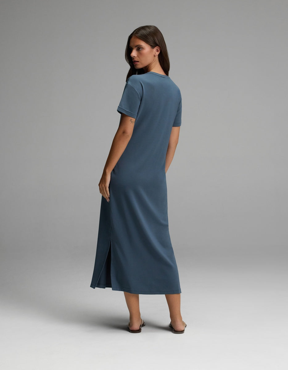 A woman stands facing away, glancing over her shoulder. She wears the Cozy Earth Women’s Brushed Bamboo Midi Dress in blue—featuring short sleeves, an ankle-length hem with a side slit—and dark sandals. The background is plain and gray.