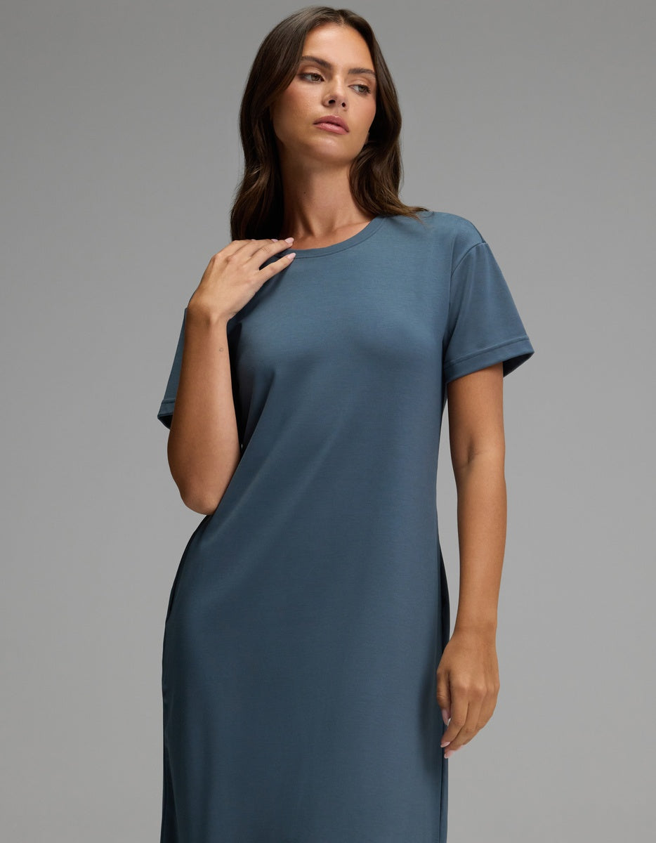 A woman with long brown hair models the Cozy Earth Women’s Brushed Bamboo Midi Dress in slate blue, featuring a loose fit and short sleeves, as she poses against a plain gray background, gazing slightly to the side with one hand gently resting near her collarbone.