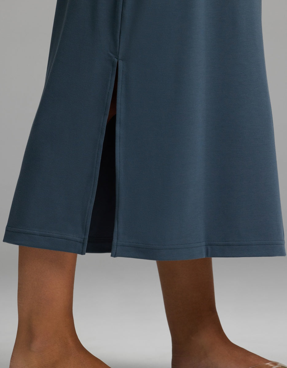 A close-up of a person’s lower legs, barefoot on a light gray surface, wearing the Cozy Earth Women’s Brushed Bamboo Midi Dress in dark blue, featuring a side slit.