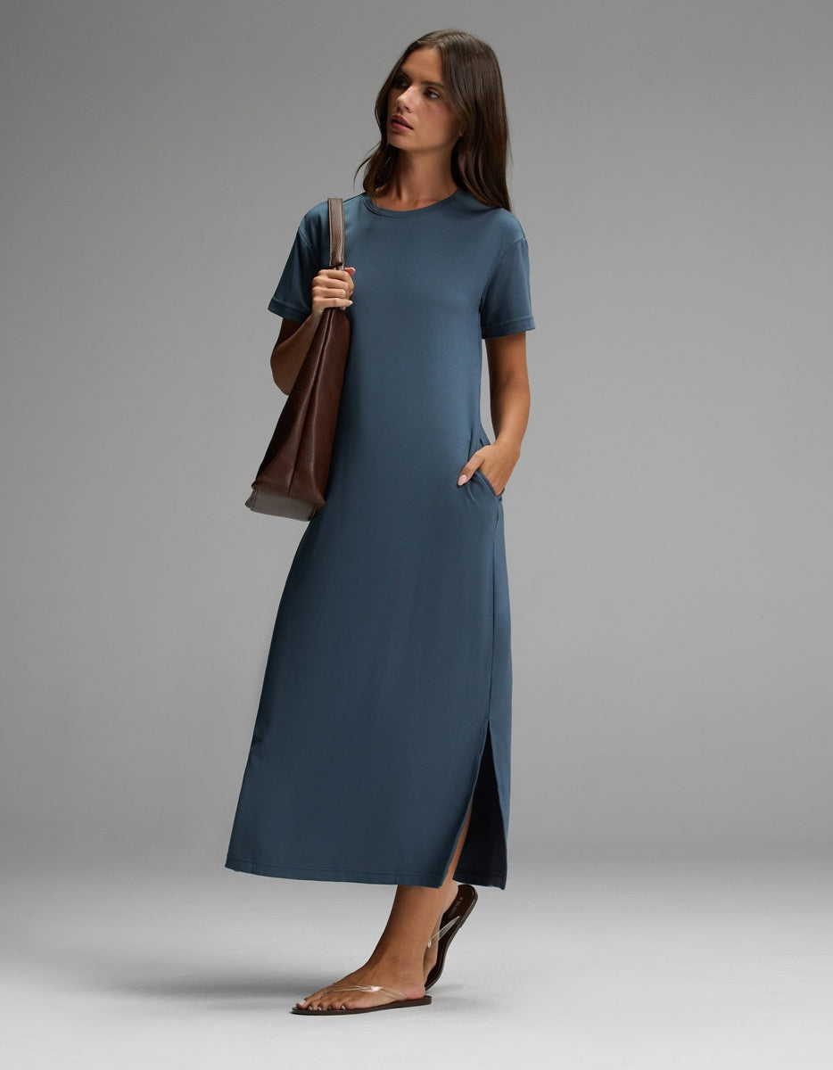 A woman stands against a plain gray background wearing the Cozy Earth Women’s Brushed Bamboo Midi Dress in teal, featuring short sleeves and an ankle-length silhouette with a side slit. She accessorizes with flat sandals and carries a large brown tote bag.