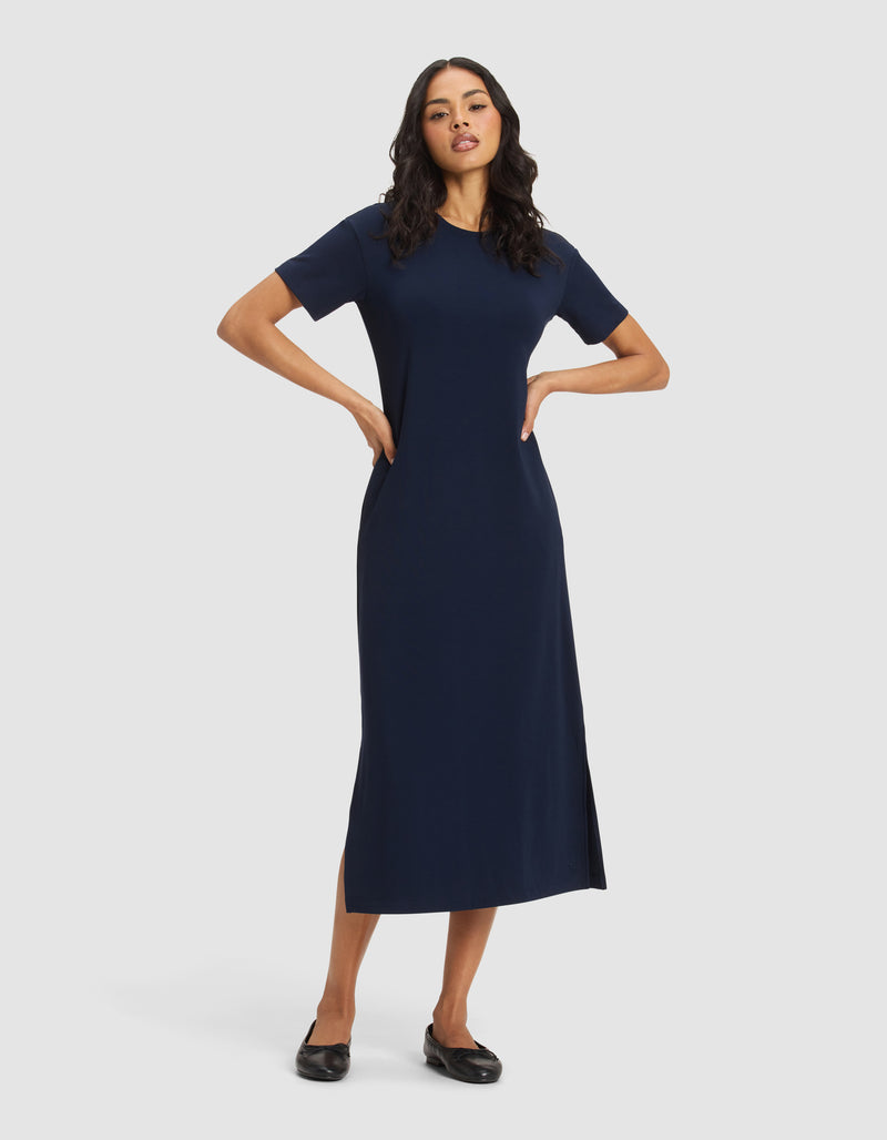 A woman with long dark hair stands against a plain background wearing Cozy Earth's Women's Brushed Bamboo Midi Dress in navy, featuring short sleeves, an ankle-length hem with side slit, and paired with black flats. Her hands rest on her hips.