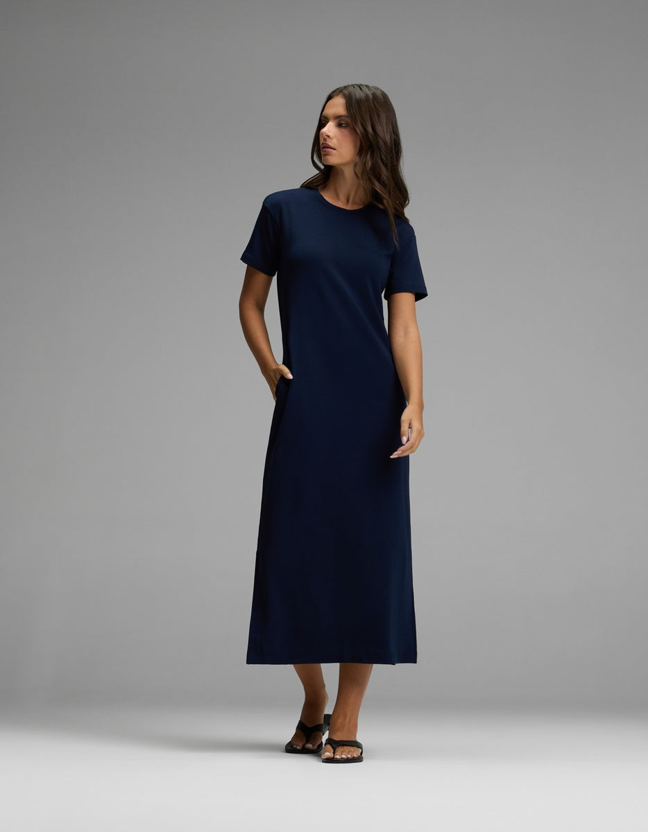 A woman stands in a studio against a plain gray background, wearing the Cozy Earth Women’s Brushed Bamboo Midi Dress in navy. The dress features short sleeves, an ankle-length hem, and convenient pockets. She has dark hair, wears sandals, and gazes slightly to the side.