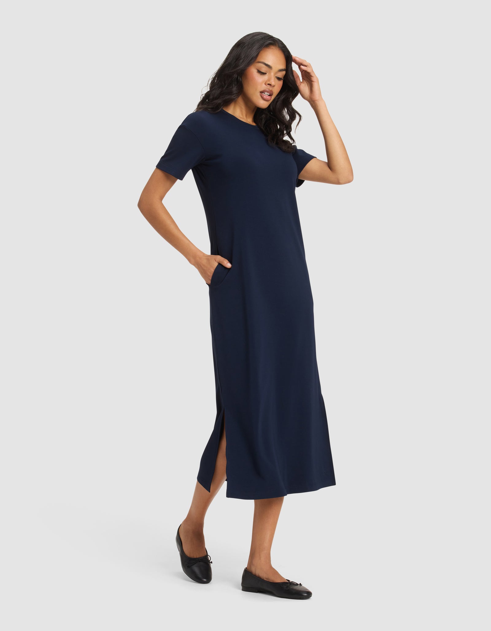 A woman wearing the Cozy Earth Women's Brushed Bamboo Midi Dress in navy blue with short sleeves and side slits, paired with black flats, stands against a light gray background, one hand in her pocket and the other touching her hair.