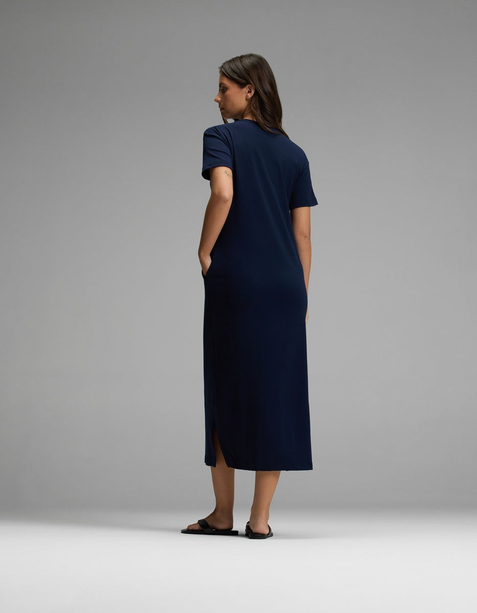 A woman stands against a plain background, facing away, wearing the Cozy Earth Women’s Brushed Bamboo Midi Dress in navy blue with short sleeves, side slits, and black sandals.