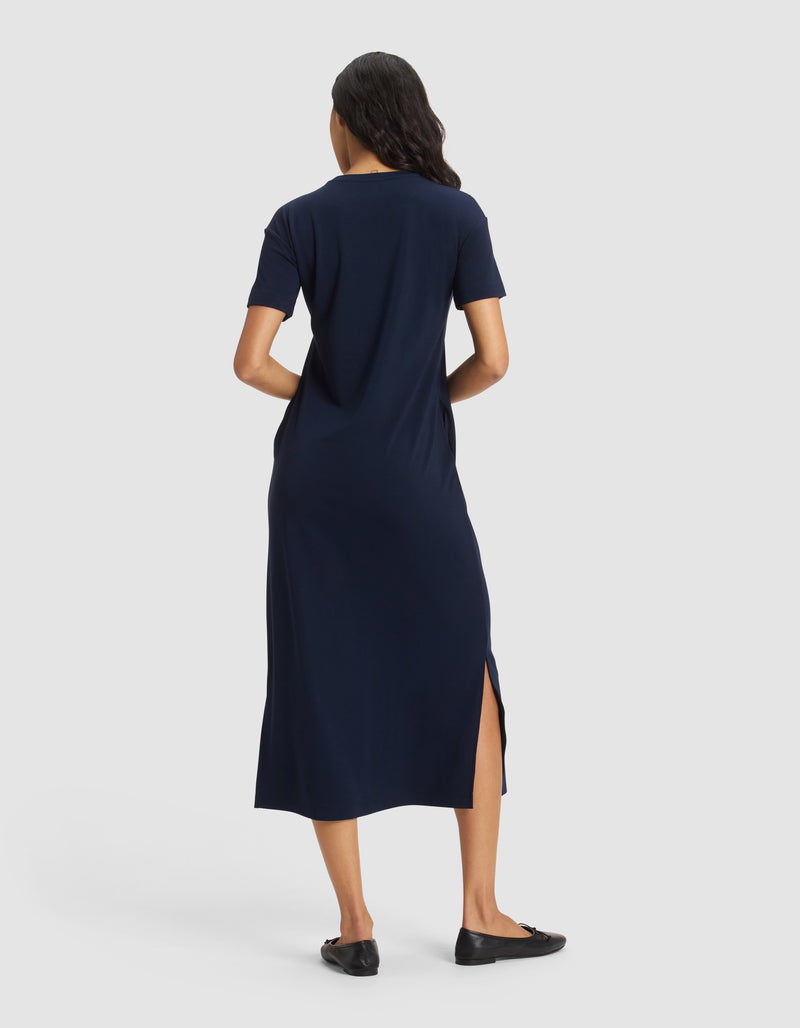 A woman with loose waves stands facing away on a plain white background, hands in her pockets, wearing Cozy Earth's Women's Brushed Bamboo Midi Dress in navy blue with short sleeves and side slit, paired with black flats.