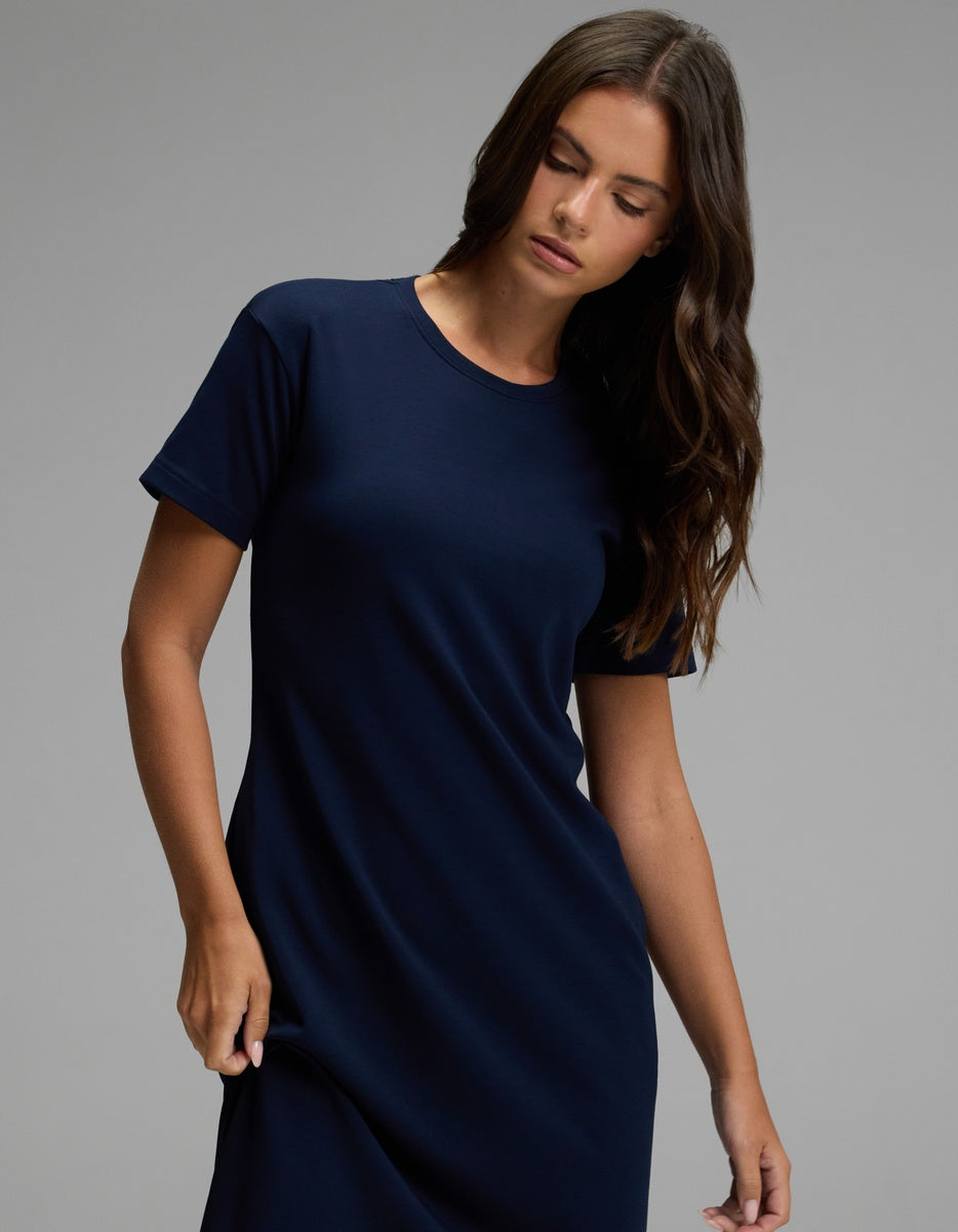 A woman with long brown hair stands against a plain gray background, looking slightly downward while holding the skirt of her Cozy Earth Women’s Brushed Bamboo Midi Dress in navy blue, which features short sleeves.