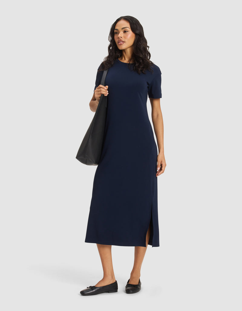 A woman wears the Cozy Earth Women's Brushed Bamboo Midi Dress in navy blue with short sleeves and a side slit, paired with black flats and a large black tote bag, standing against a plain light background.