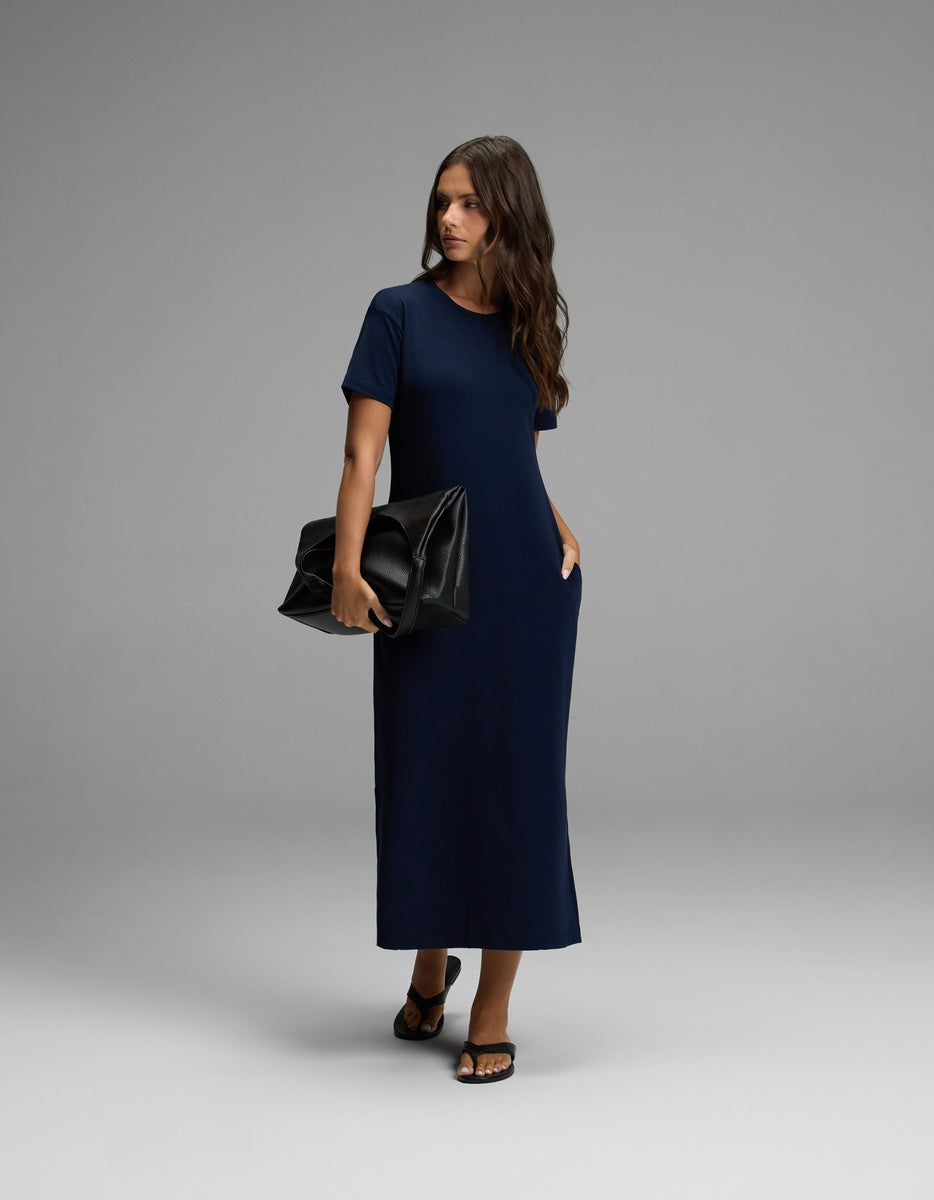 A woman wearing the Cozy Earth Women’s Brushed Bamboo Midi Dress in navy blue, paired with black sandals and holding a large black clutch, stands against a plain gray background.