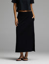A woman wearing Cozy Earth’s Women’s Brushed Bamboo Midi Skirt in black, paired with a black crop top and black sandals, stands with one hand in her skirt pocket against a plain gray background.|Color:Black