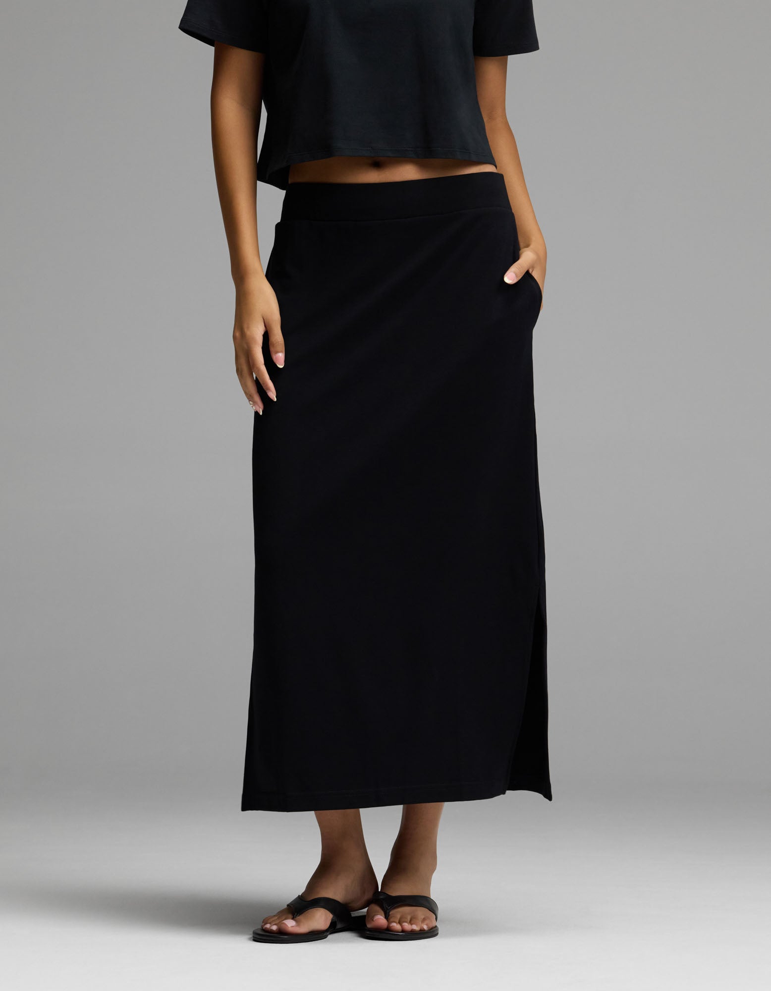 A woman wearing Cozy Earth’s Women’s Brushed Bamboo Midi Skirt in black, paired with a black crop top and black sandals, stands with one hand in her skirt pocket against a plain gray background.