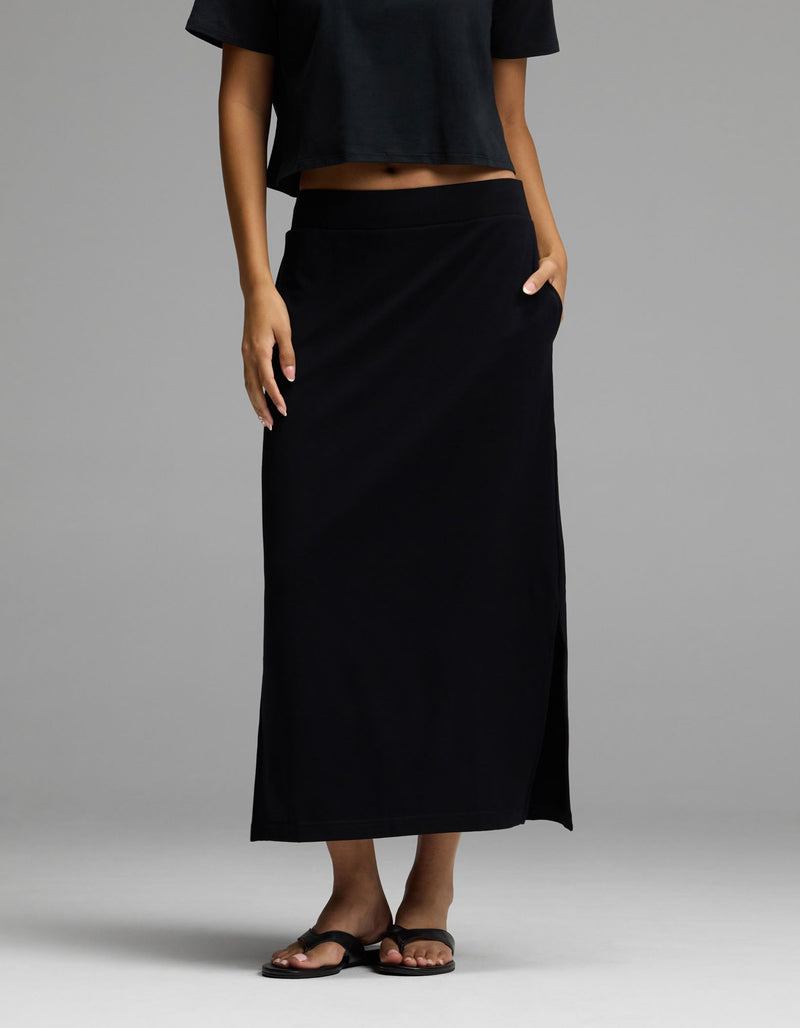 A woman wearing Cozy Earth’s Women’s Brushed Bamboo Midi Skirt in black, paired with a black crop top and black sandals, stands with one hand in her skirt pocket against a plain gray background.