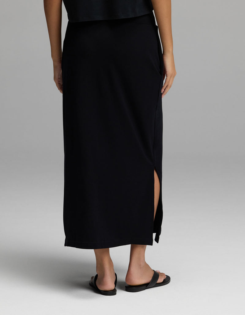 A person is shown from the back, wearing Cozy Earth’s Women’s Brushed Bamboo Midi Skirt in black—with a side slit—paired with black sandals, standing against a light gray background. Their upper body is partially visible.