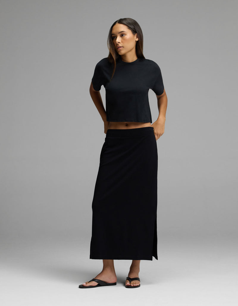 A woman stands in a studio wearing the Cozy Earth Women’s Brushed Bamboo Midi Skirt in black, paired with a black short-sleeve crop top and black sandals against a neutral background.