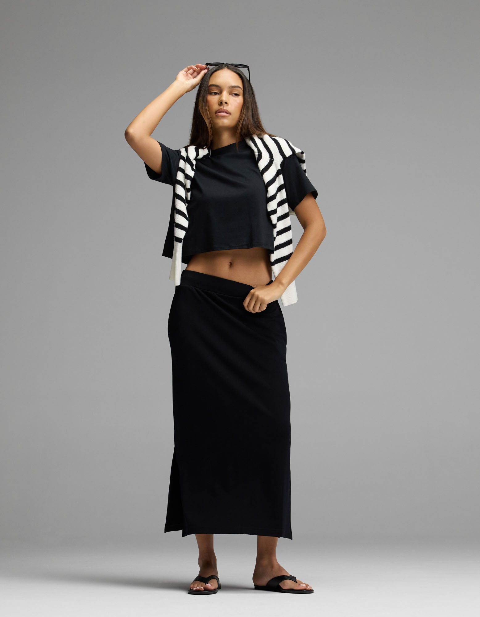 A woman stands barefoot against a plain gray background, wearing the Cozy Earth Women’s Brushed Bamboo Midi Skirt in black with a matching black crop top. Draped over her shoulders is a black and white striped sweater, and she holds sunglasses on her head.