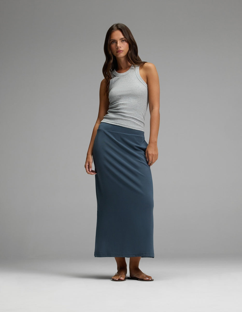 A woman stands against a plain gray background, wearing a light gray sleeveless top, sandals, and the Cozy Earth Women’s Brushed Bamboo Midi Skirt in dark blue. She has long, wavy hair and a neutral expression.