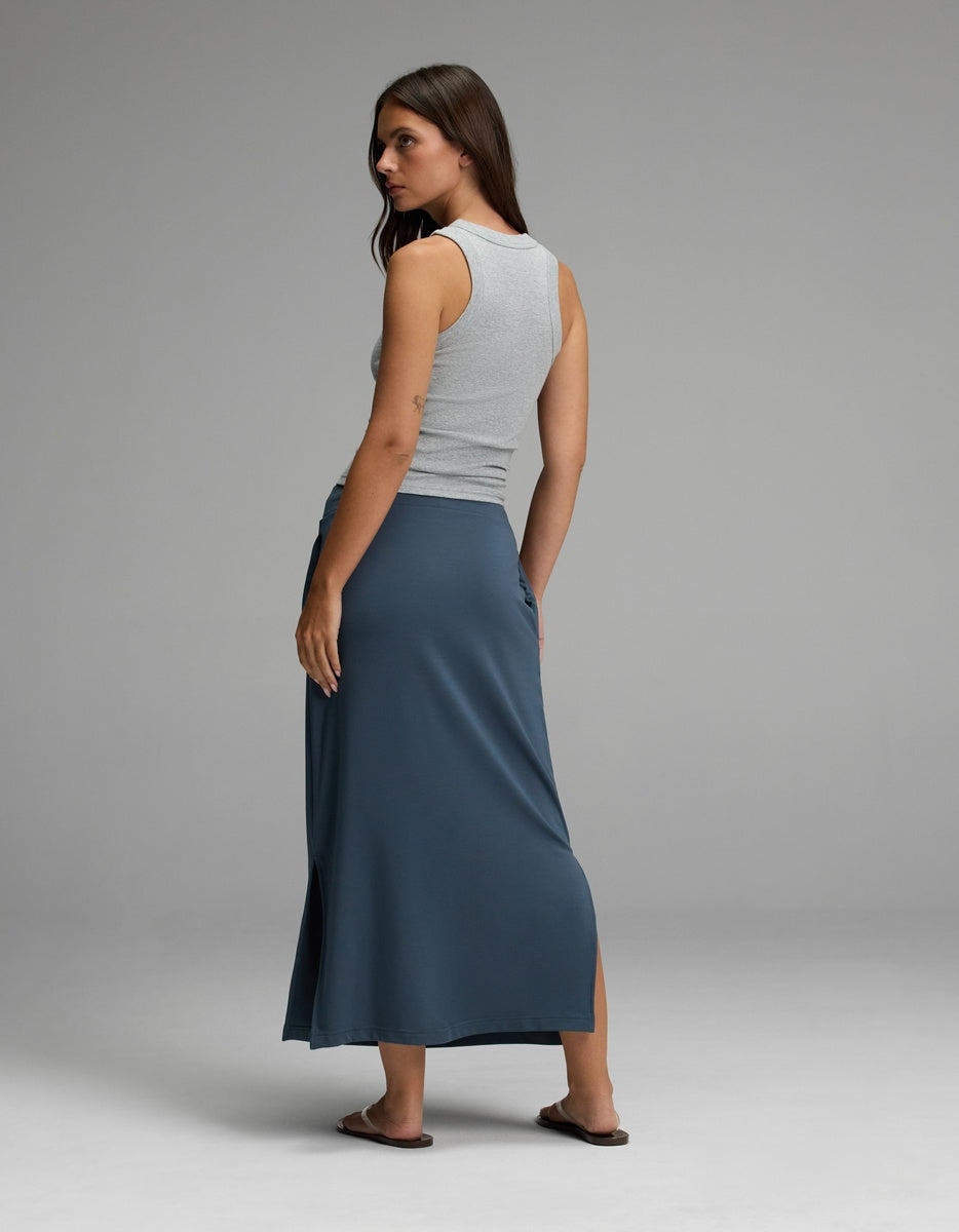 A woman stands with her back to the camera, wearing a sleeveless gray top and the Cozy Earth Women’s Brushed Bamboo Midi Skirt in blue, featuring a side slit. She pairs the outfit with sandals and glances over her shoulder against a plain gray background.