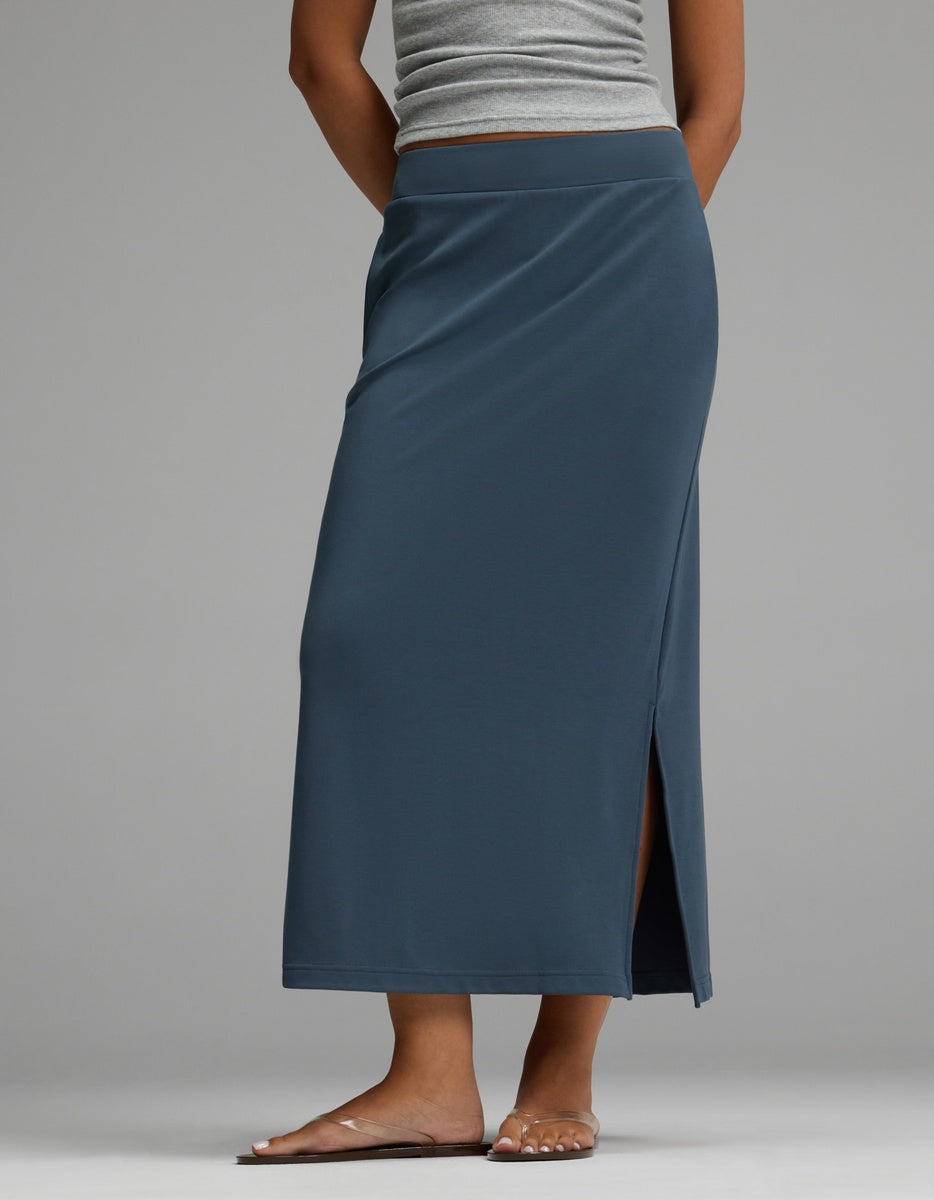 A person is shown from the waist down, barefoot, wearing a Cozy Earth Women’s Brushed Bamboo Midi Skirt in blue with a side slit, paired with a gray tank top, standing against a light gray background.
