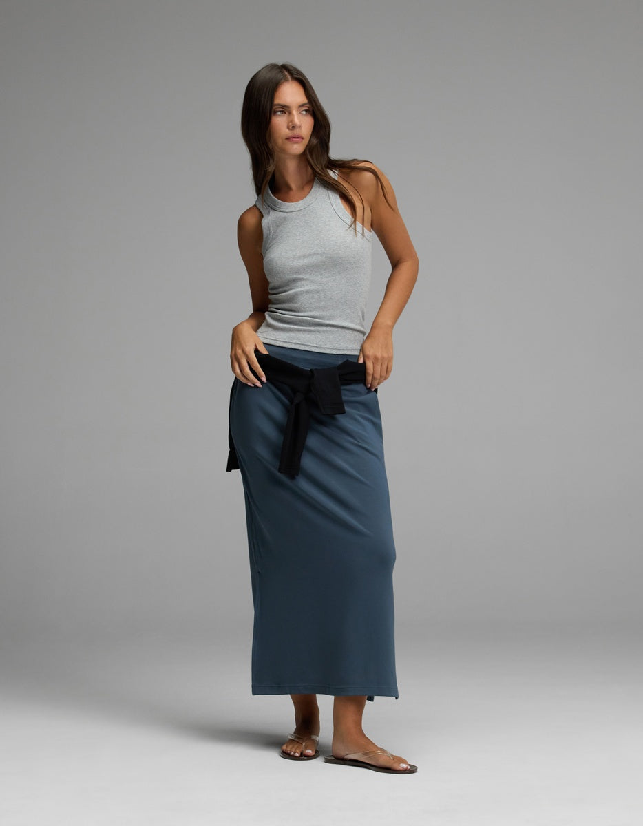 A woman poses confidently against a simple gray backdrop, wearing the Cozy Earth Women’s Brushed Bamboo Midi Skirt in blue paired with a light gray tank top, a black sweater tied at her waist, and sandals. Her long brown hair adds to her relaxed look.