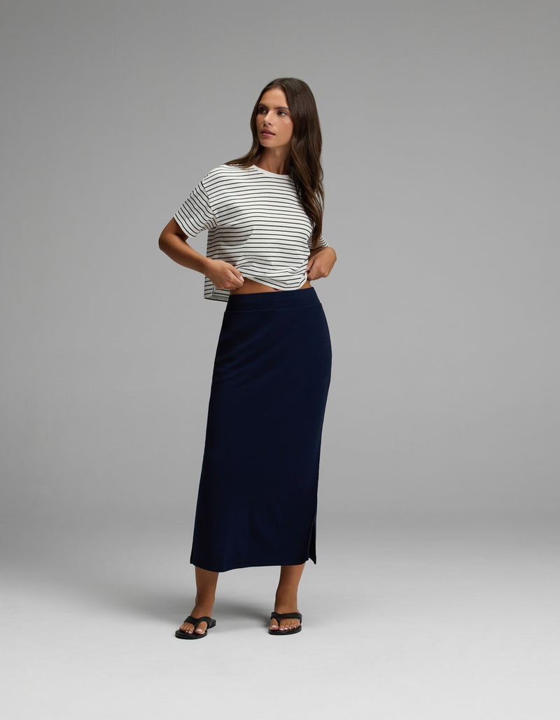 A woman stands in a studio wearing a black and white striped t-shirt, the Cozy Earth Women’s Brushed Bamboo Midi Skirt in navy with a side slit, and black sandals. She looks to the side with one hand adjusting her shirt.